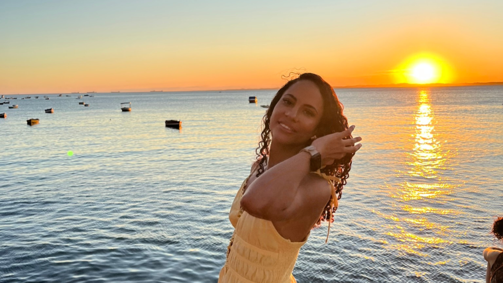 Jade Sande apreciando o pôr do sol no MAM Salvador, ao lado da Praia da Gamboa, com o céu alaranjado refletindo no mar, um destaque do turismo em Salvador Bahia.